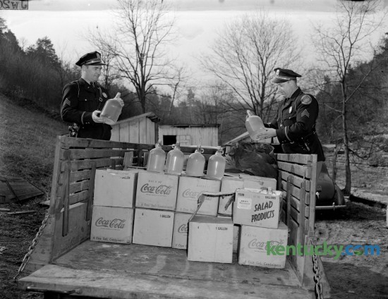Two State Highway Patrol officers posed with some of the confiscated moonshine after Federal agents raided a the production facility at Fox Gap on March 14, 1945. The location of the still was on the side of a creek near the old Fox Gap school on the Frankfort-Lawrenceburg pike, three miles from Frankfort. Officials stopped this truck, loaded with 176 gallons of whiskey, as it was on it's way to Lawrenceburg.  Four suspects were taken into custody. Published in the Lexington Herald March 15, 1945. Herald-Leader Archive Photo